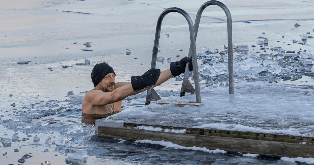 Man ice swimming next to a frozen dock after sauna, holding onto a metal ladder in icy water